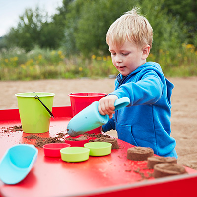 Un jeune garçon joue avec des outils en sable sur une table de jeu dans une cour de récréation.